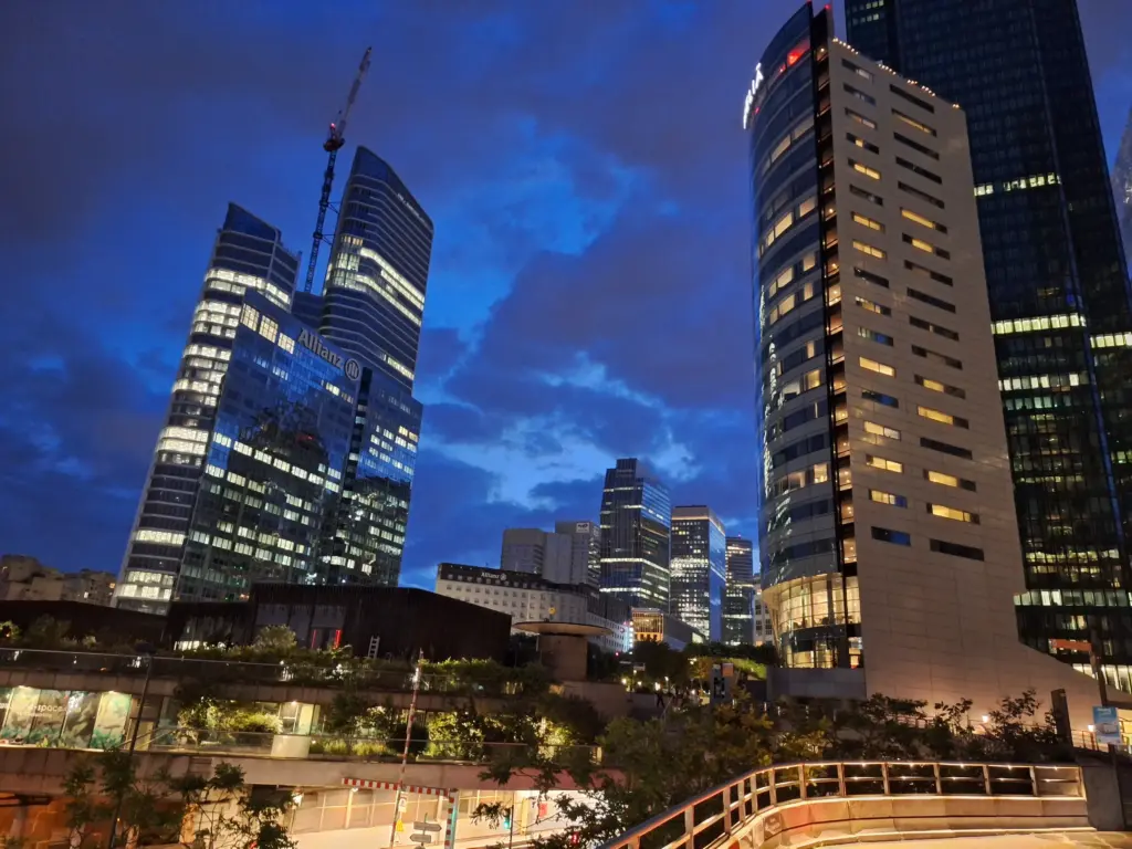 Blick auf die Hochhäuser des Pariser Geschäftsviertels La Défense bei Nacht, mit beleuchteten Fassaden und dramatischem Himmel